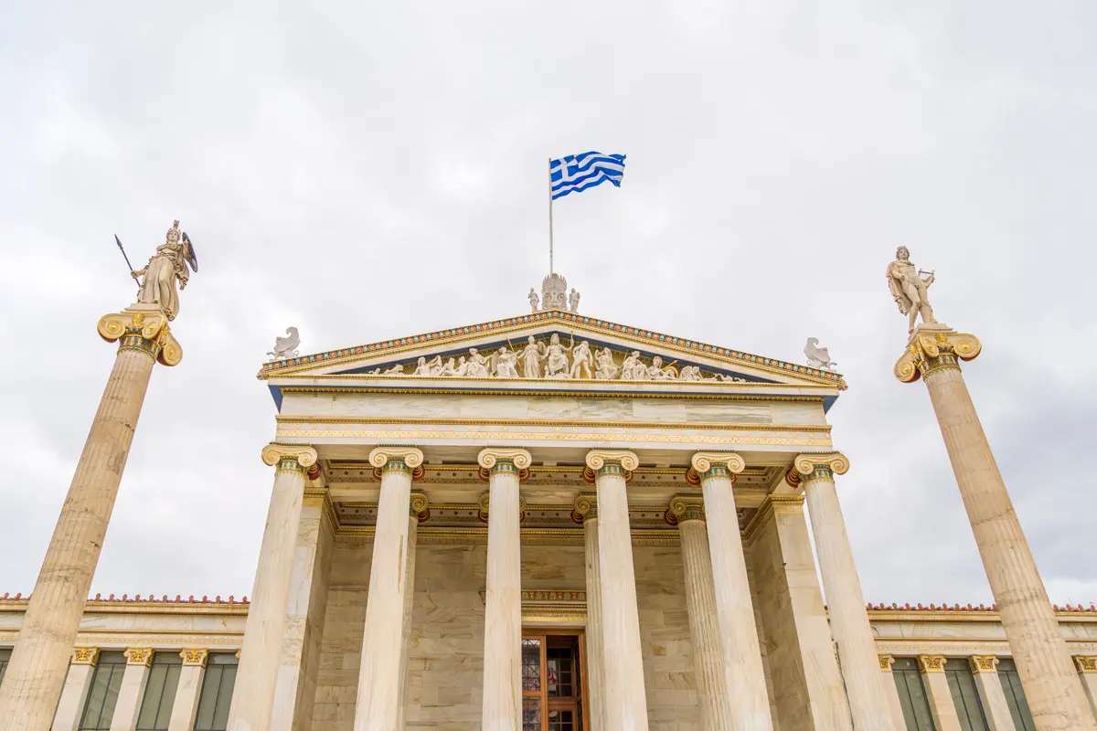 The National Academy of Athens tands in the daylight with a bright sky overhead, a Greek flag waves proudly from the roof.