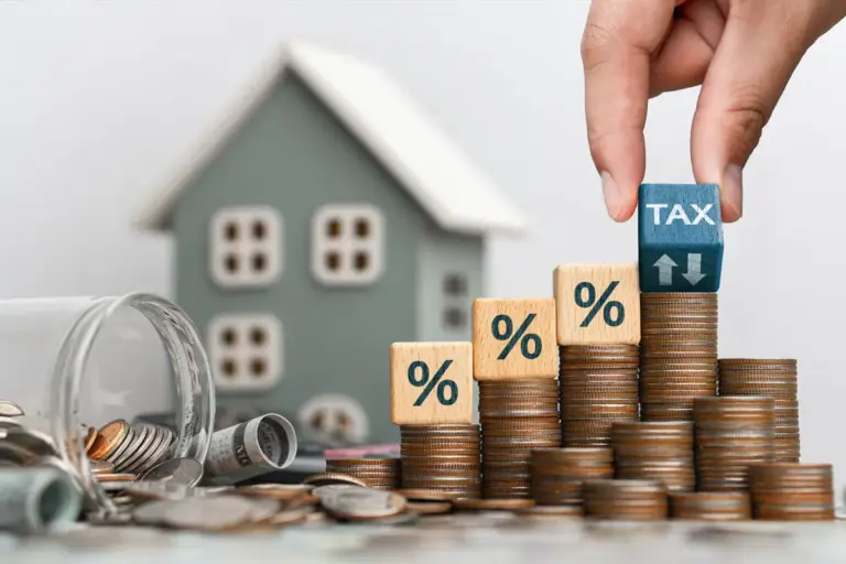 A hand places a cube with the word "TAX" on top of a stack of coins near a model house, illustrating a property tax concept.