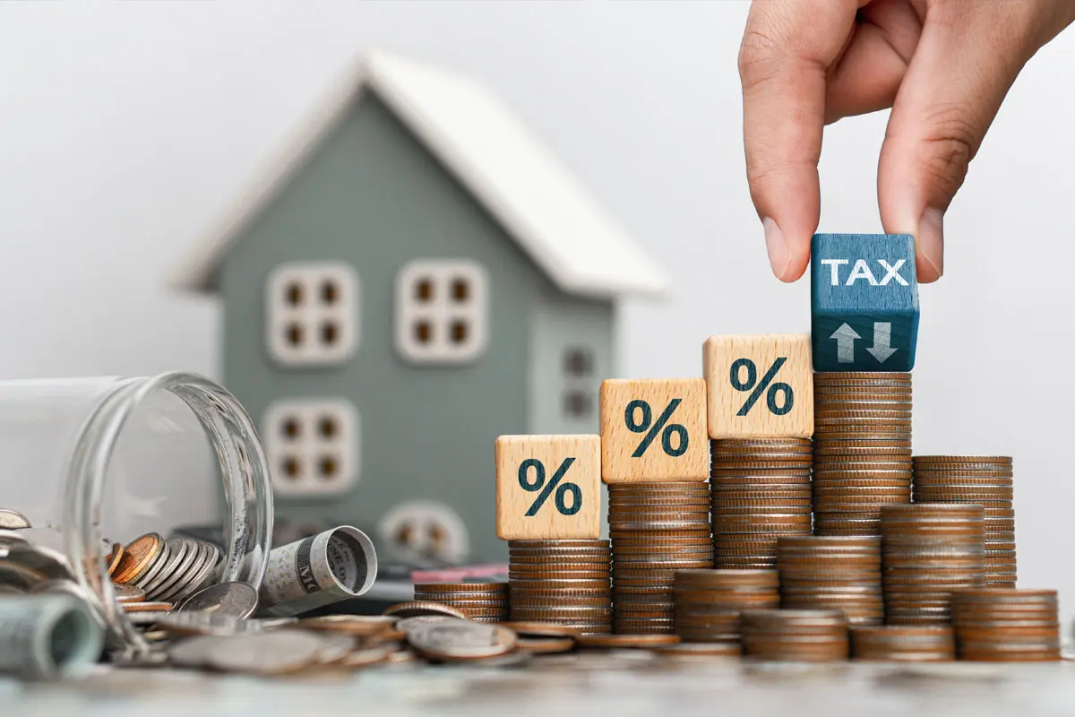 A hand places a cube with the word "TAX" on top of a stack of coins near a model house, illustrating a property tax concept.